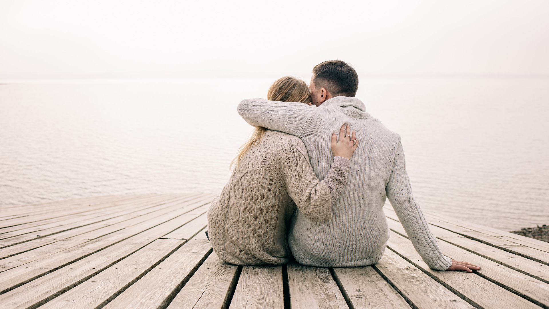 Blonde woman and brunette man dressed in beige and grey cardigans sitting down next to a pier and hugging each other.