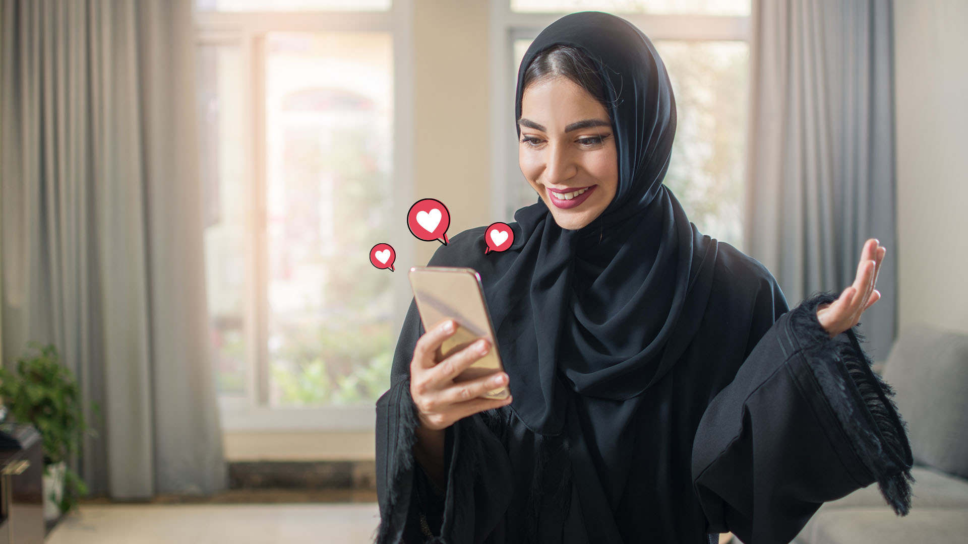 Woman dressed in black with black hijab, holding her phone in her hand and smiling while looking at it. She is in living room