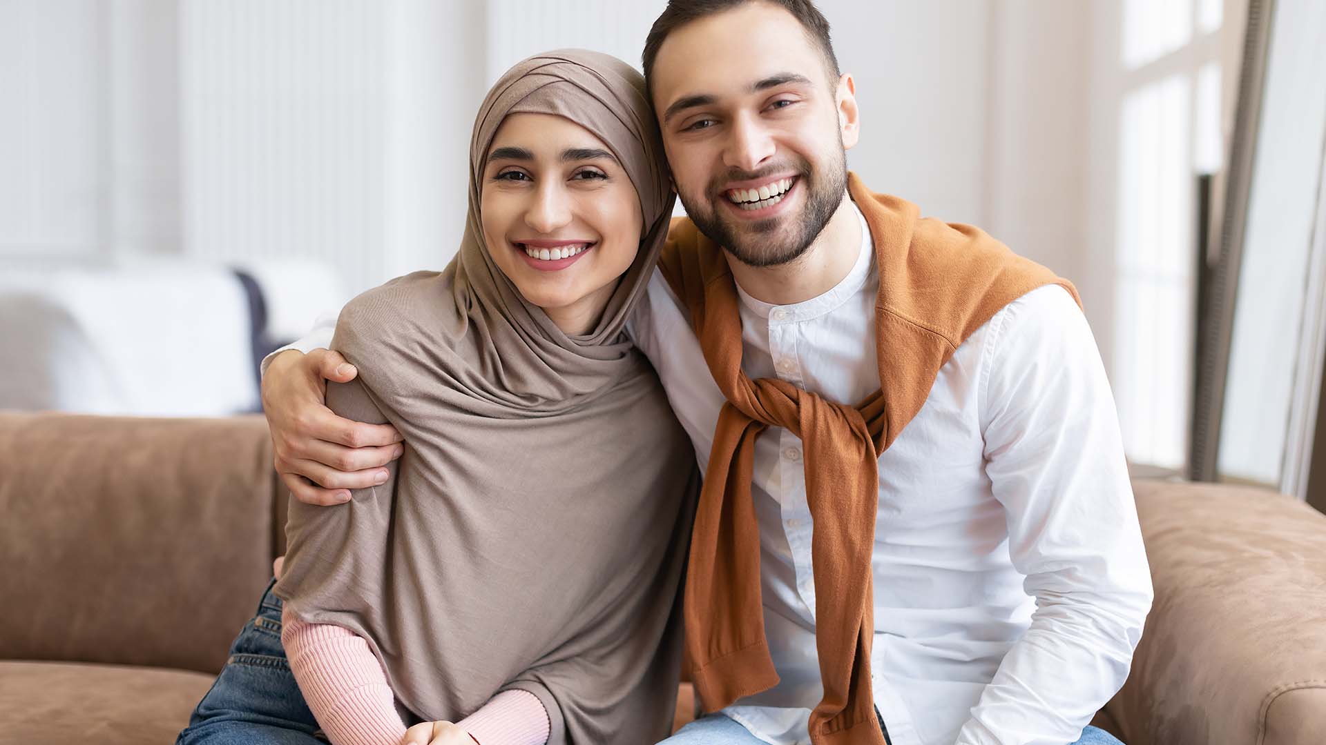 Brunette man with 5 o'clock shadow hugging a woman wearing a grey hijab next to him. Both are smiling while sitting on couch.