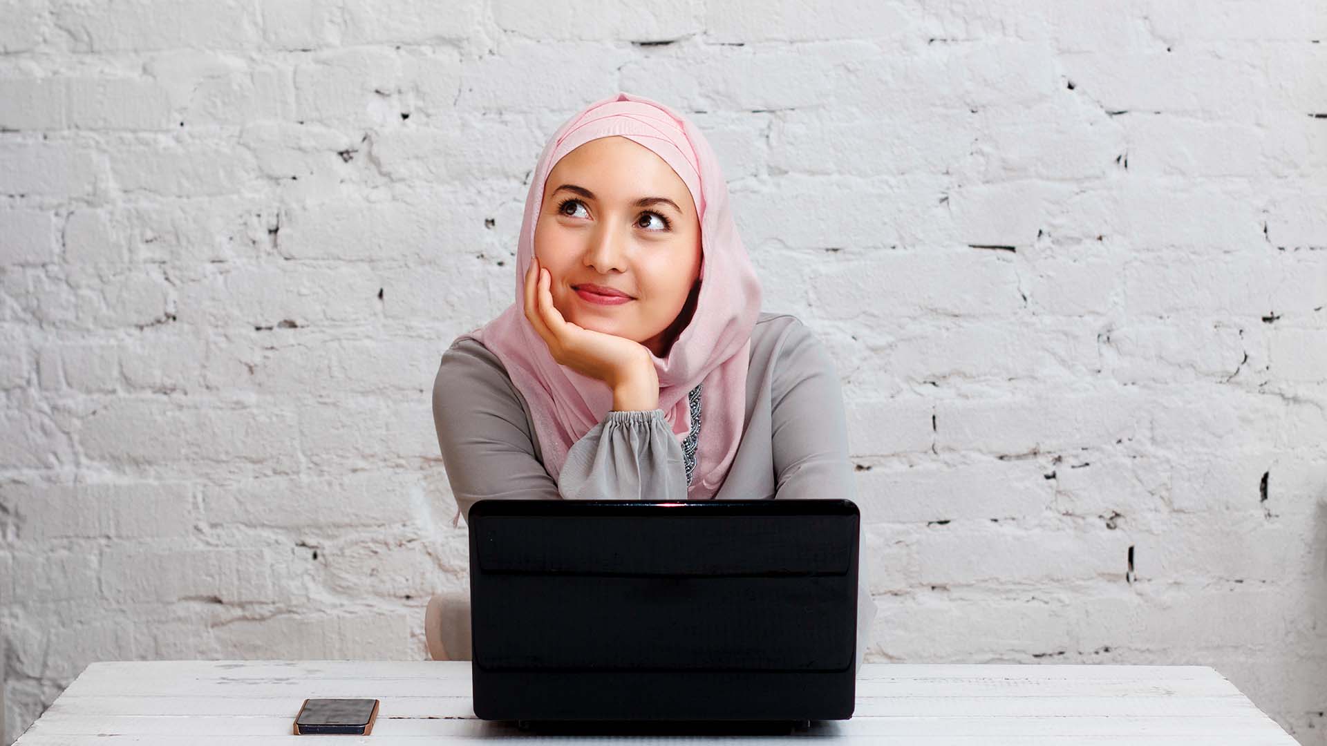 Woman wearing a pink hijab resting head on palm, with laptop and phone in front of her, thinking. White brick wall background
