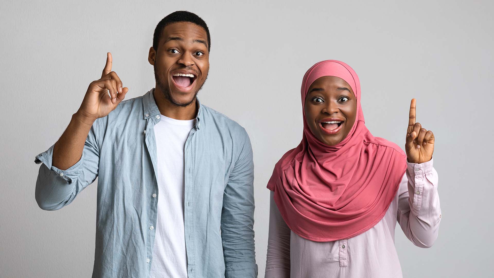 Brunette man in light blue shirt and woman in dark pink hijab holding their index fingers in the air and smiling. White wall.