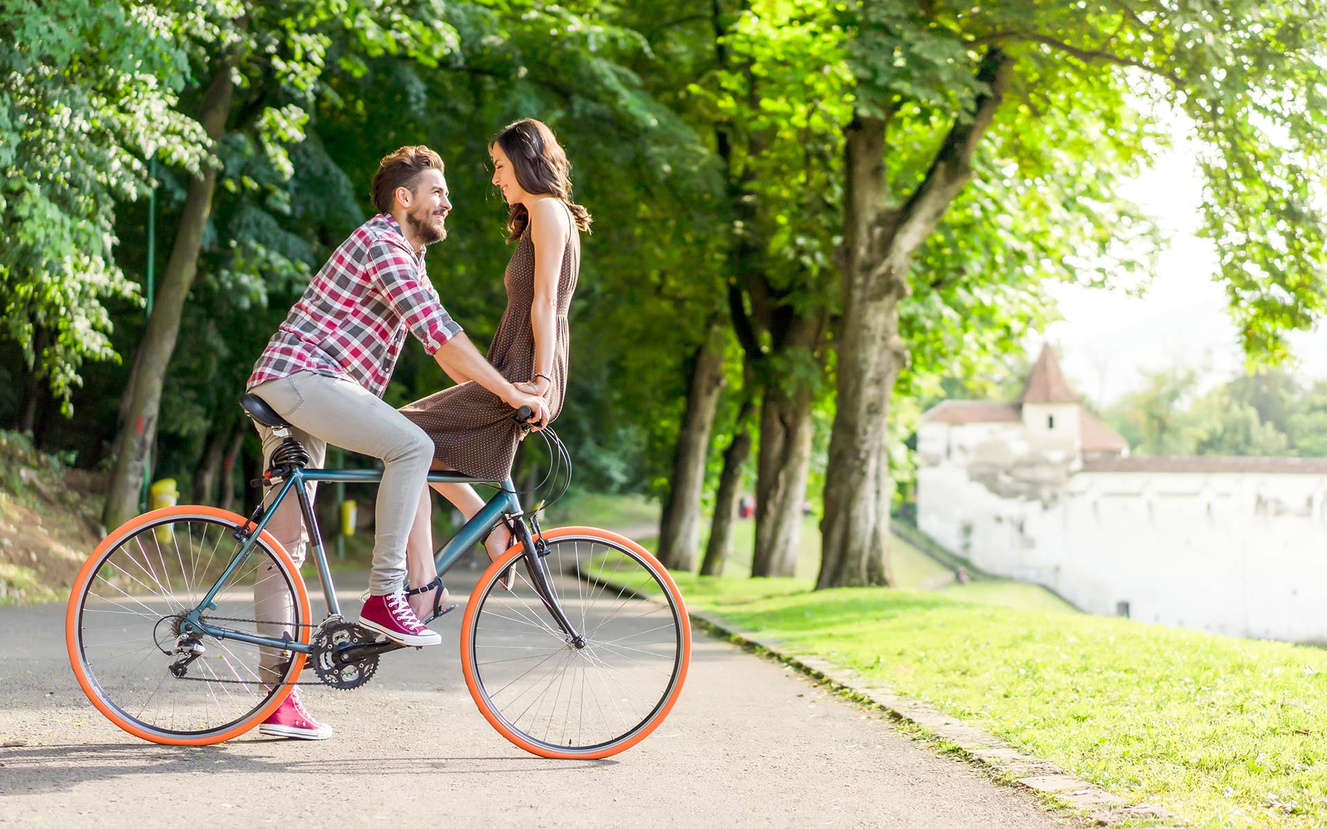 Brunette man with a beard riding a bicycle with brunette woman in brown dress sitting on the wheel looking at him.