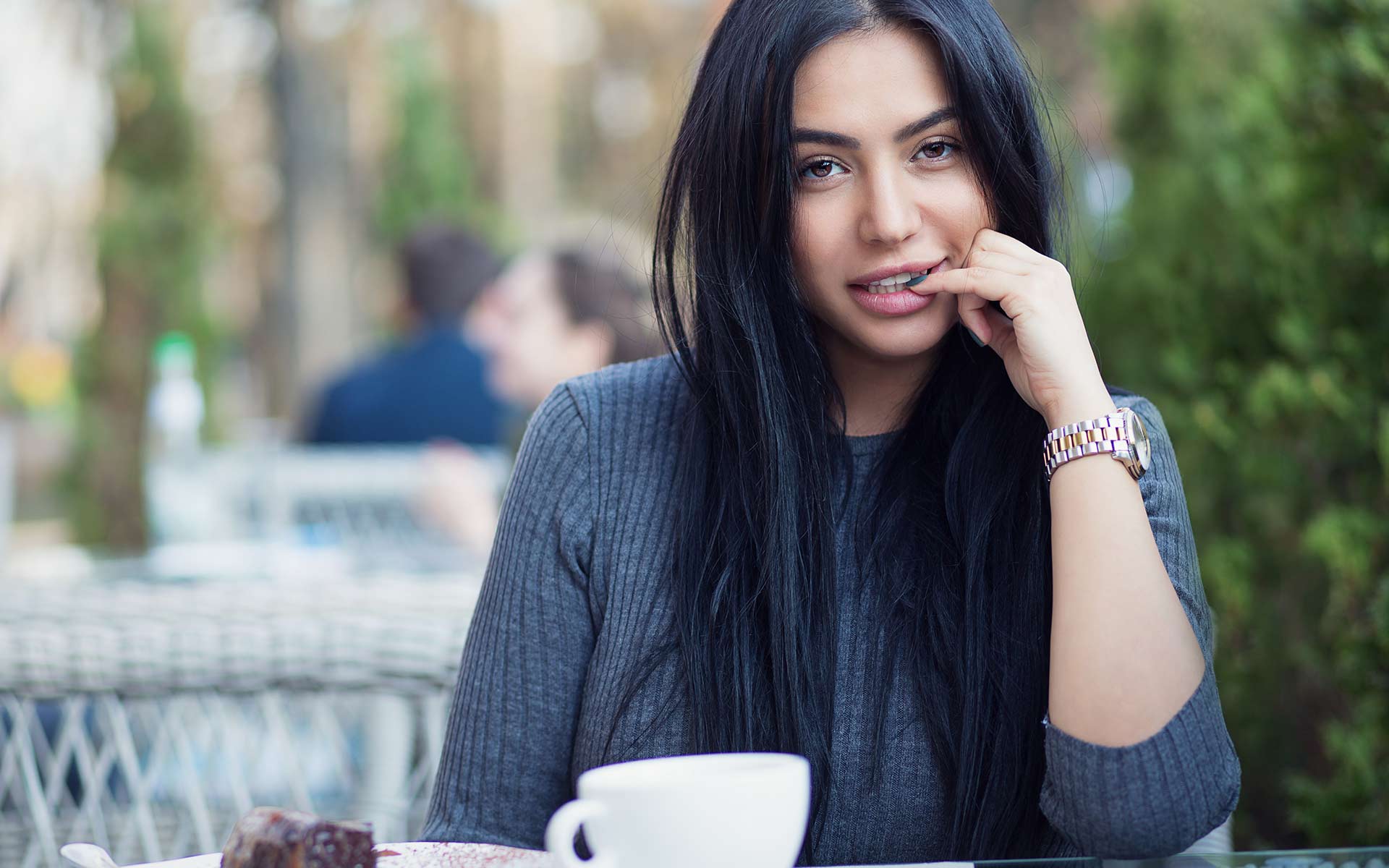 Woman in grey blouse biting the nail of her pinky finger while smiling. She has a coffee mug and cake in front of her.