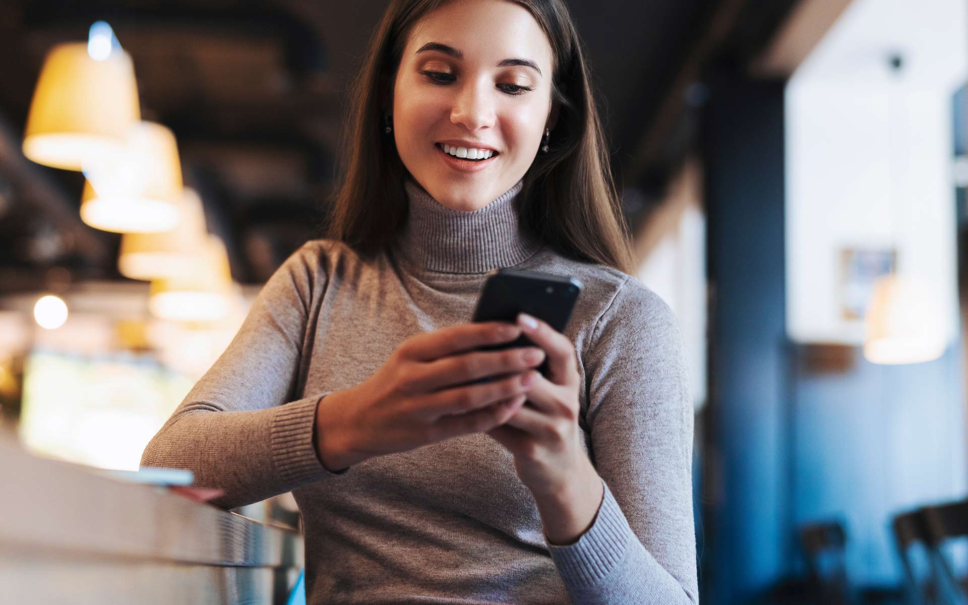 Brunette woman in grey turtleneck holding her phone with both hands and looking down on it while smiling.