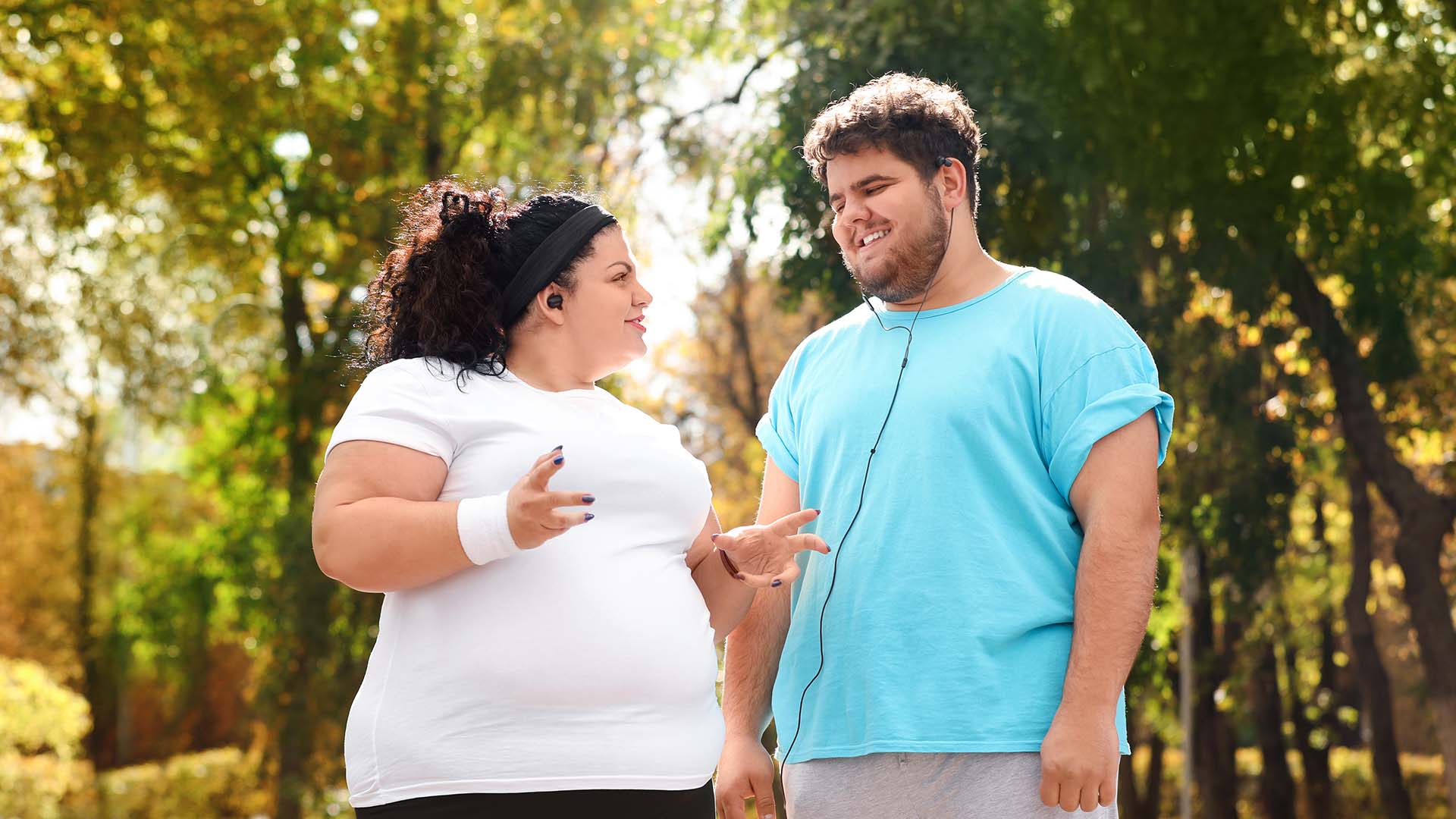 Plus size brunette man and woman standing next to each other in workout clothes, talking while smiling.