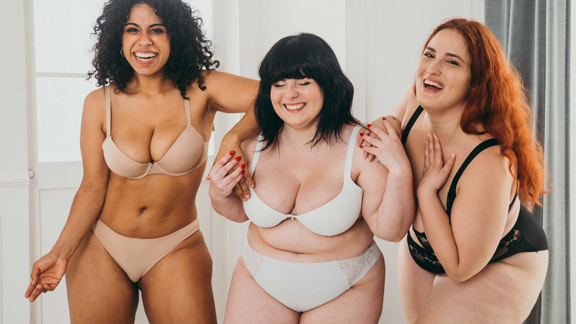 three plus-sized women in their underwear standing next to each other and laughing. Grey curtain in the background.