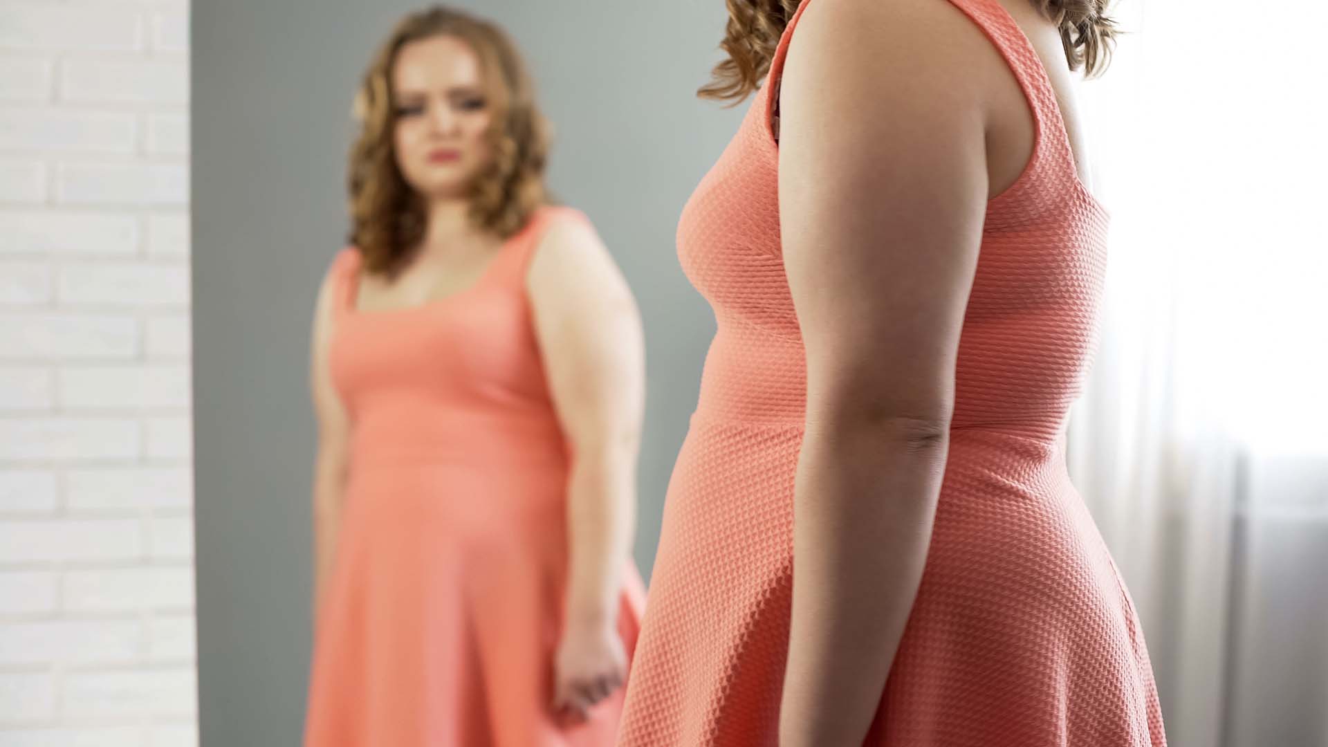Blonde BBW in light coral dress standing in front of the mirror and looking sad. Window in the background.