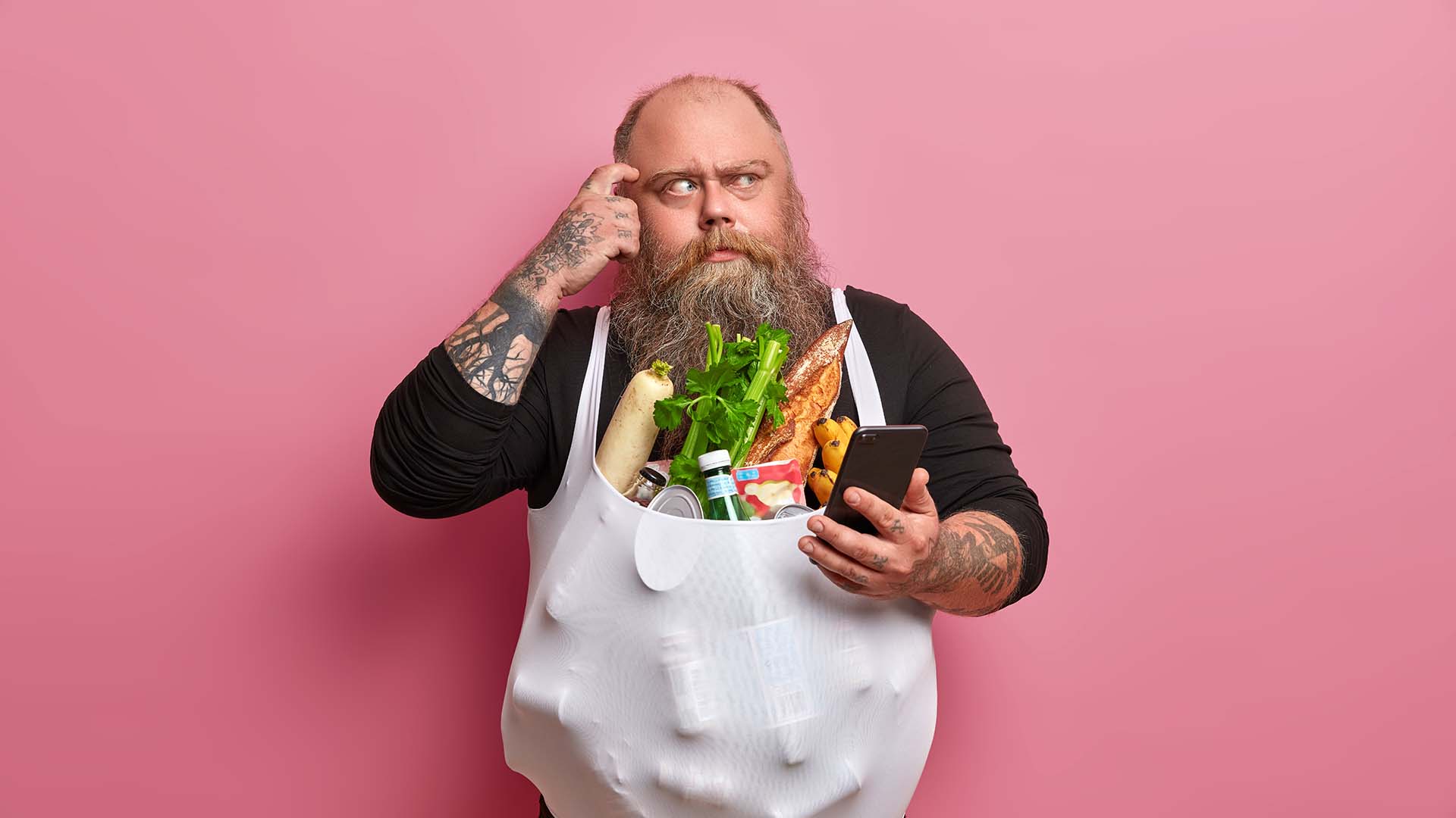 Plus size man in full beard holding his phone and looking confused. Several vegetables attached to the front of his shirt.