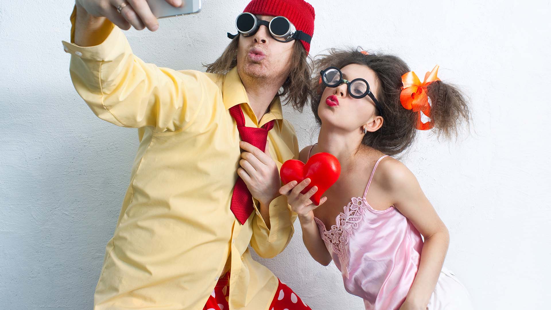 Brunette man and woman taking a selfie. The man is dressed in a yellow and red suit and woman in pink silk pajamas.