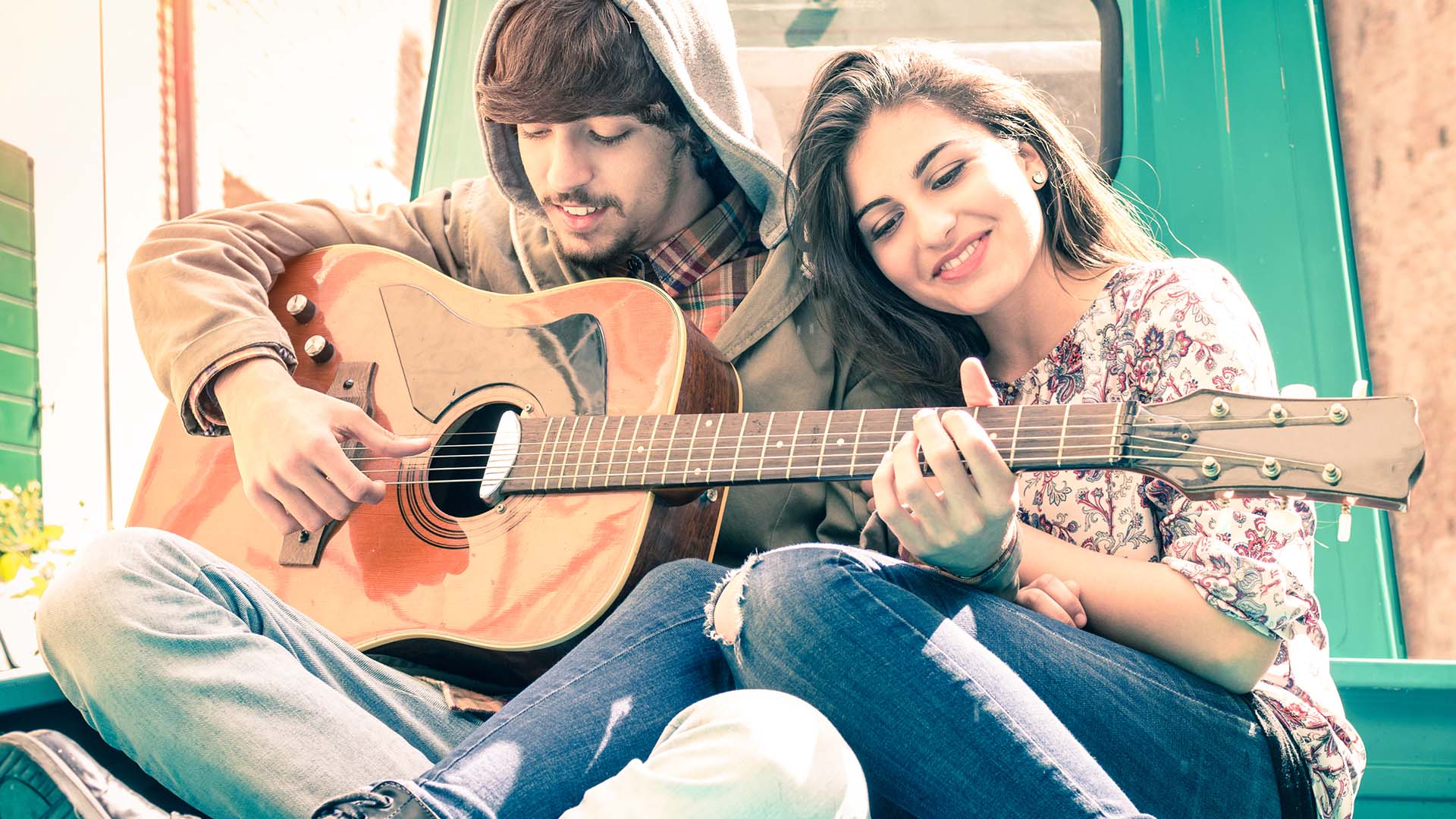 Brunette man in hoodie playing guitar while brunette woman in floral shirt sits next to him resting her head on his shoulder.