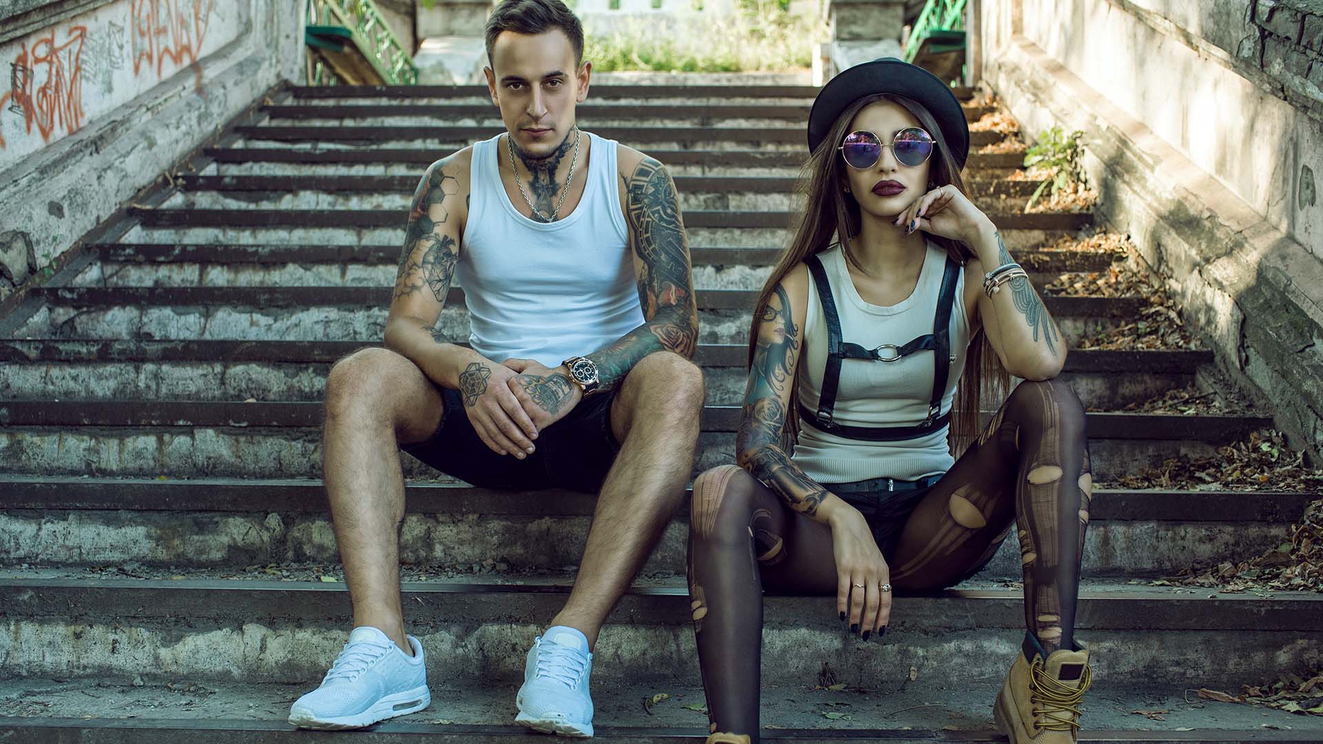 Man and woman sitting on stairs in public with multiple tattoos on their body. The woman is wearing ripped pantyhose.