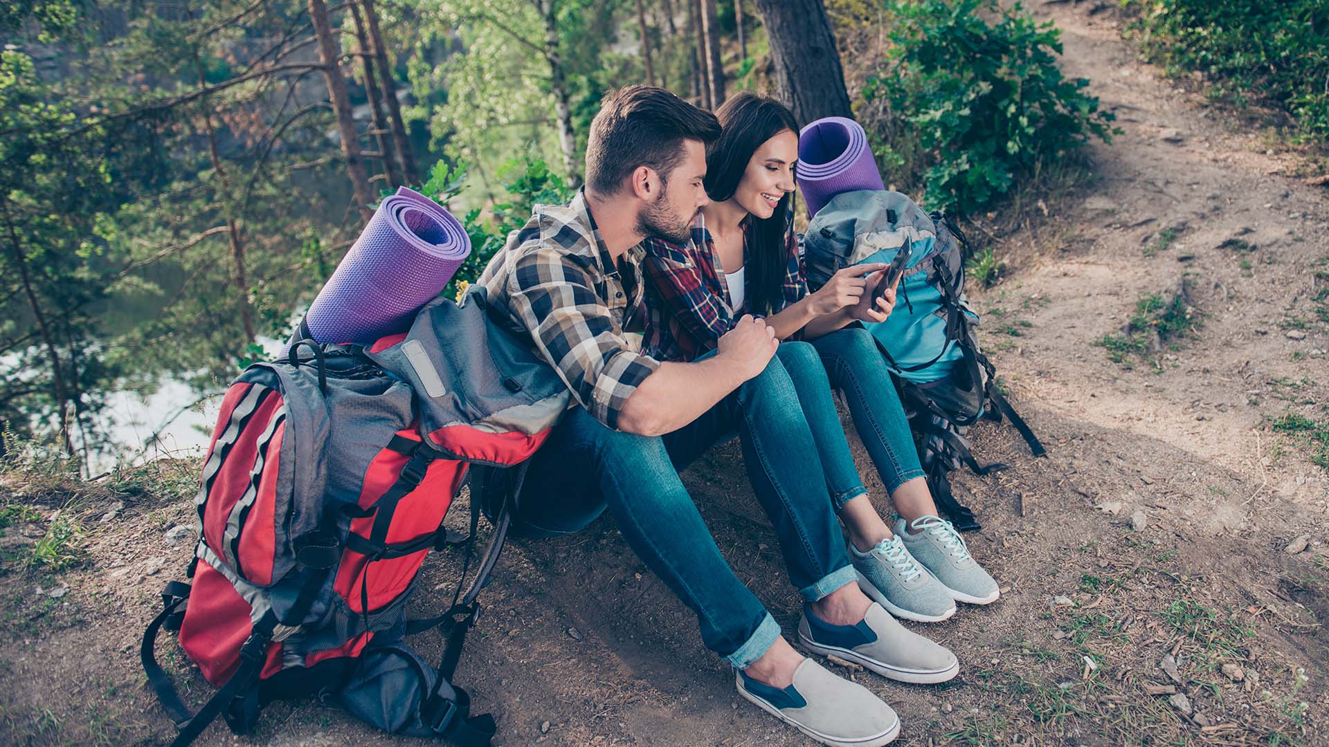 Brunette man and woman in checkered shirts with hike backpacks, sitting on the side of a nature trail looking at a phone.