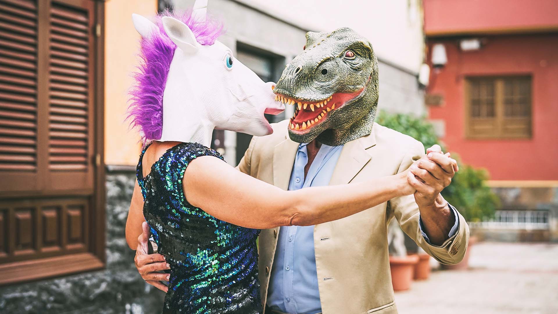 Woman in unicorn mask and man in crocodile mask slow dancing outside of a red house's porch. There are plant pots behind them