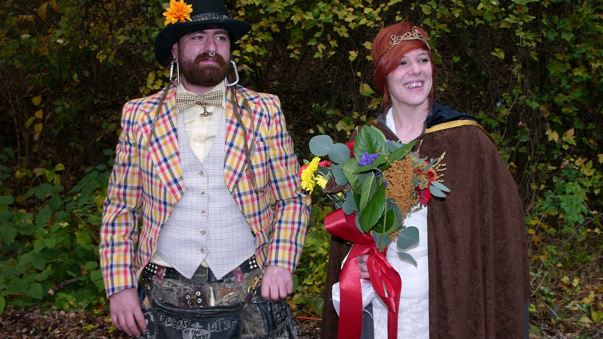 Brunette man and red-head woman dressed in outdated clothing and posing for a picture. The woman is holding flowers.