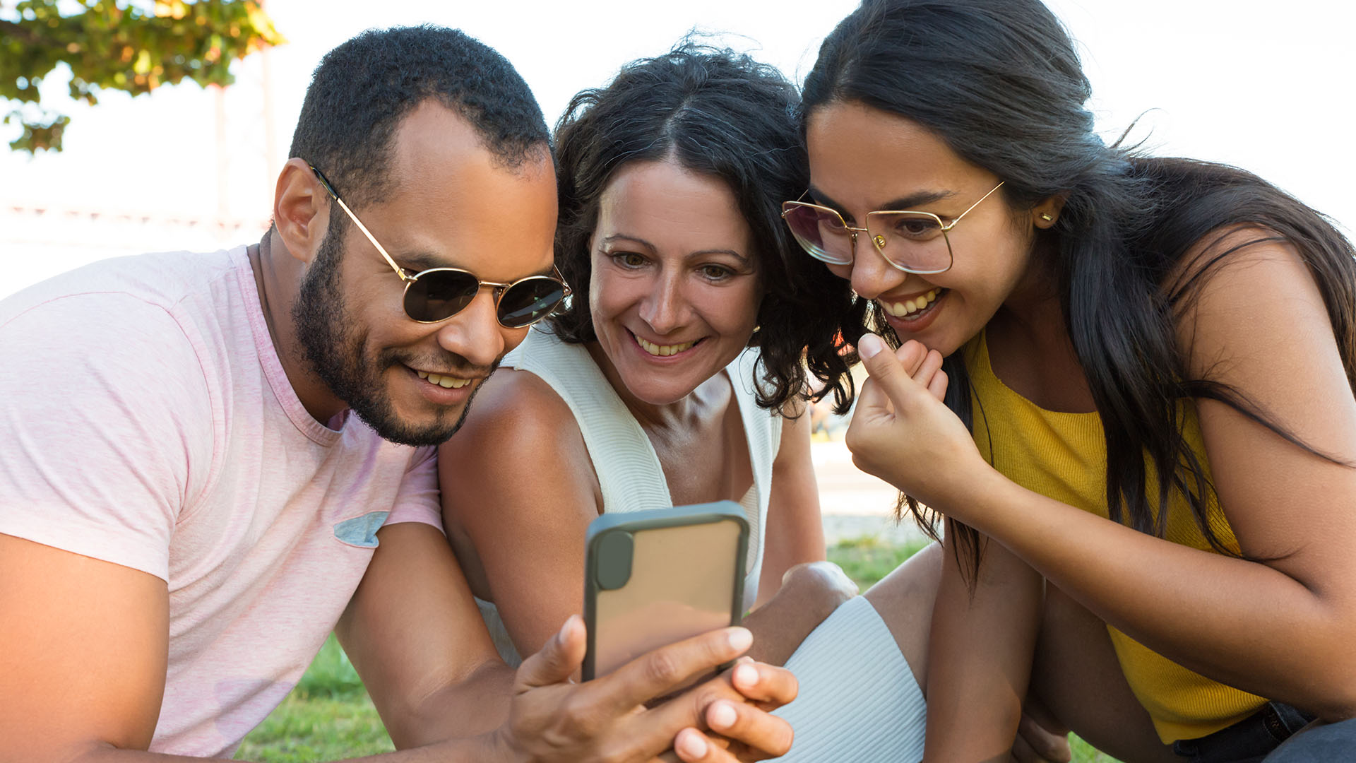 Brunette man and two women from different ethnicities, smiling while looking at at a phone. They are at a park.
