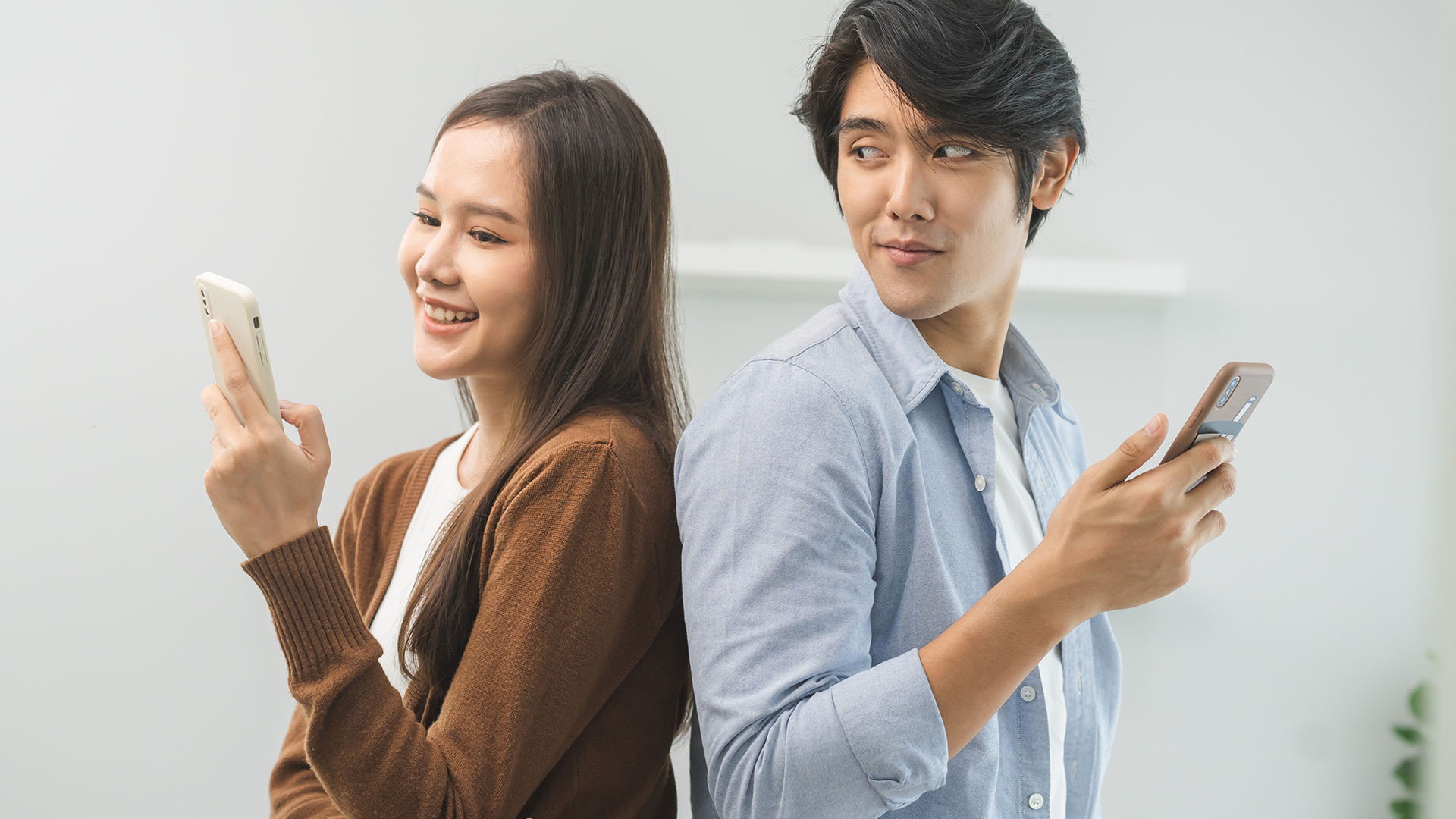 Brunette Asian man and woman, standing back to back and holding their phones. The man is looking at the woman who is smiling.