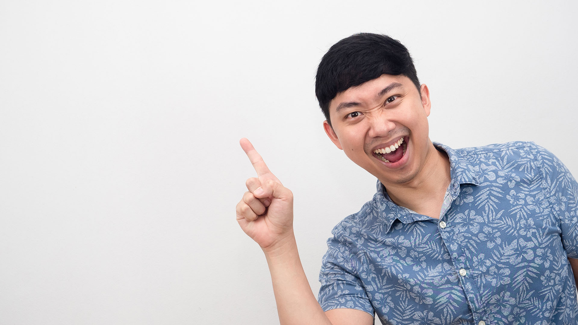 Brunette asian man in flower-print blue shirt, pointing his index finger upwards and smiling. White background.