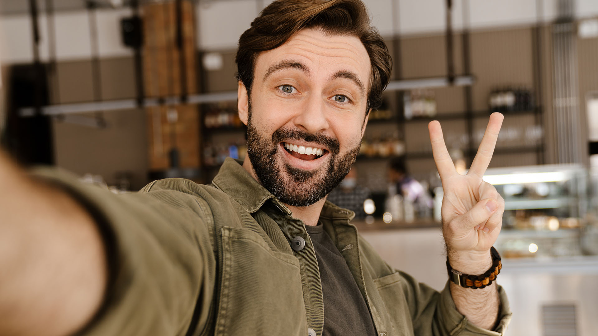 Brunette man with beard, giving the piece sign to the camera with one hand while holding it with the other. Blurry background