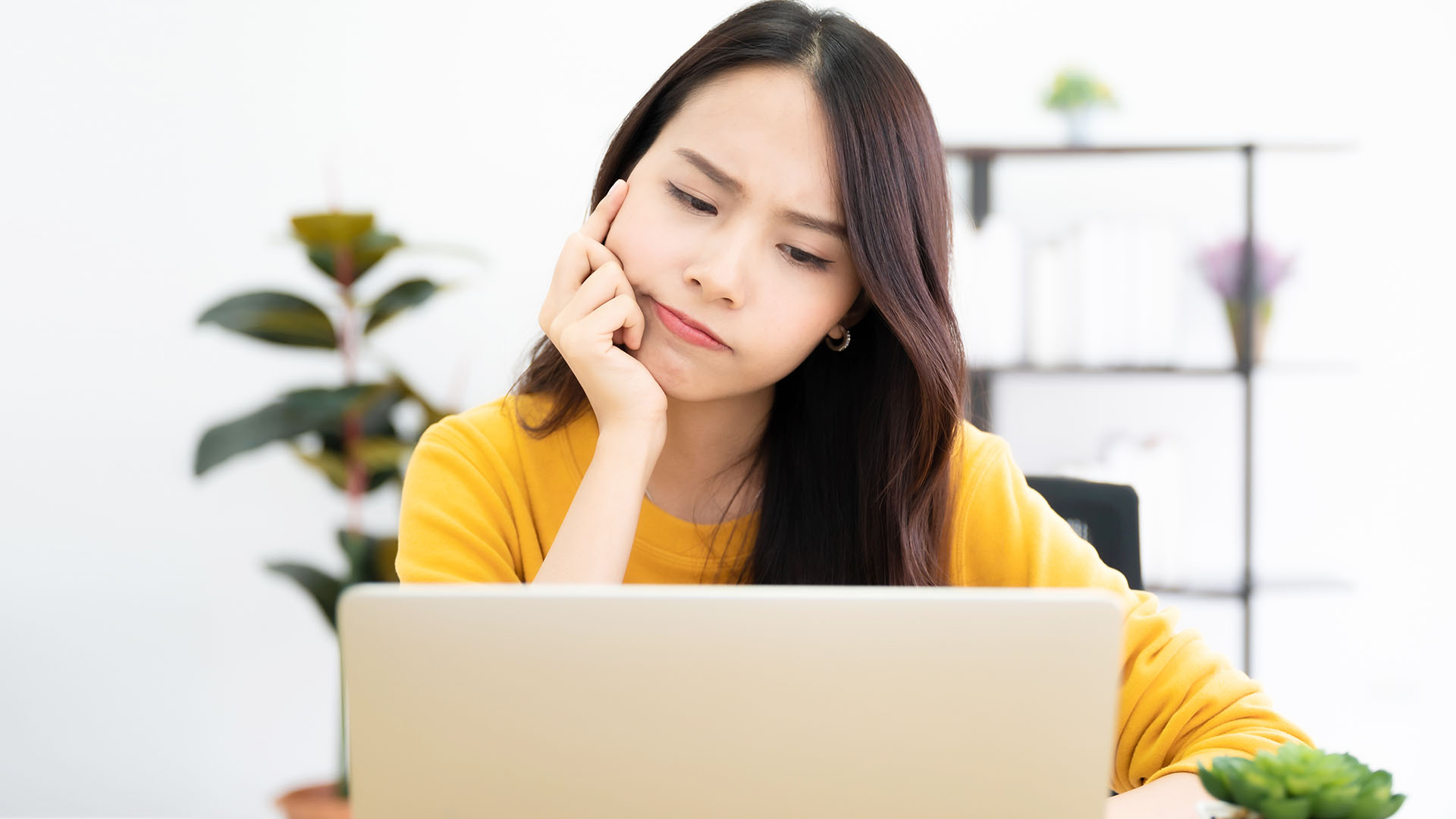 Brunette Asian woman in yellow long-sleeved shirt, sitting at desk and looking at her laptop confused. Plants in background.