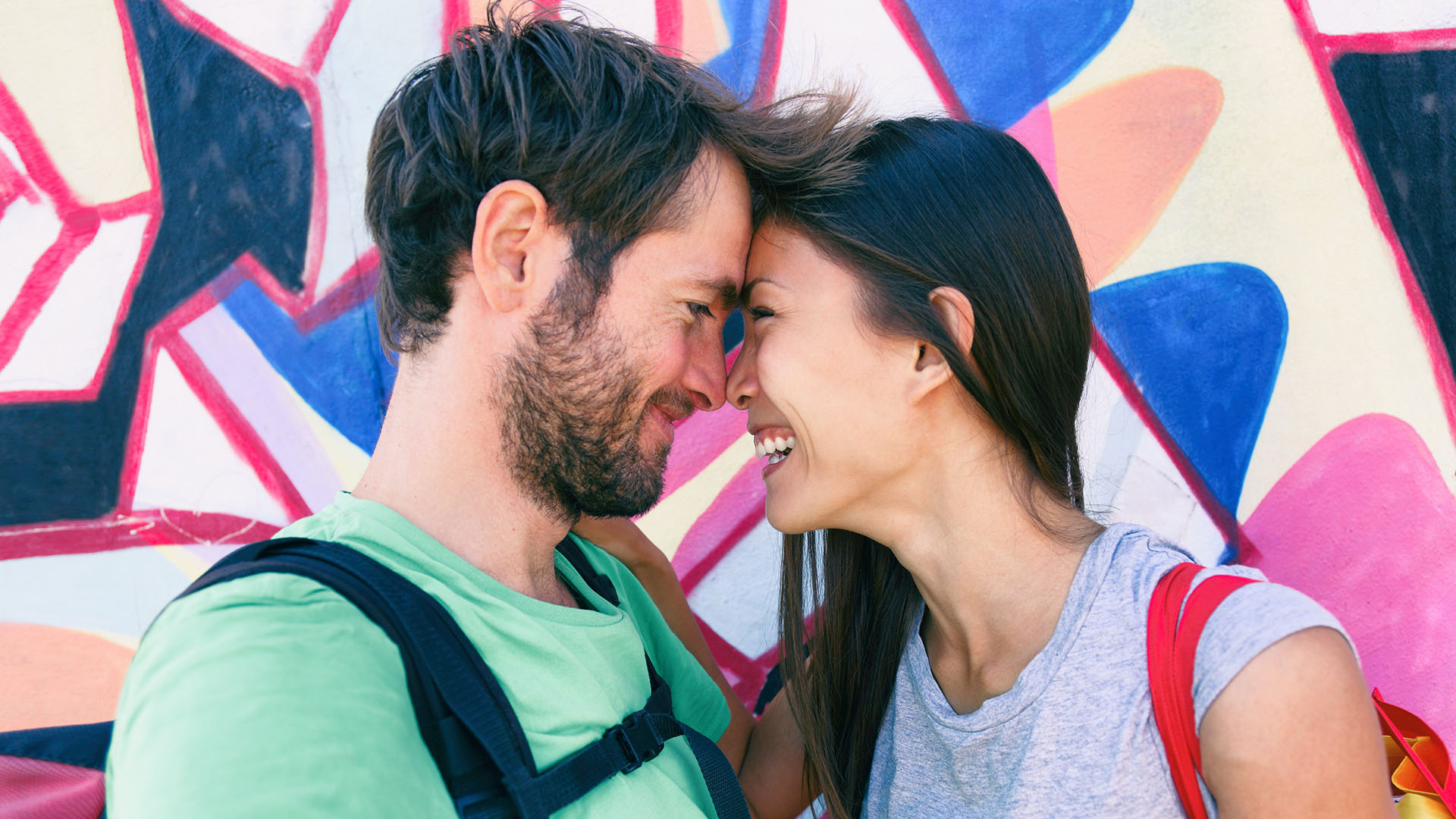 Brunette Asian woman and man in front of graffiti wall, touching their foreheads together while laughing together.