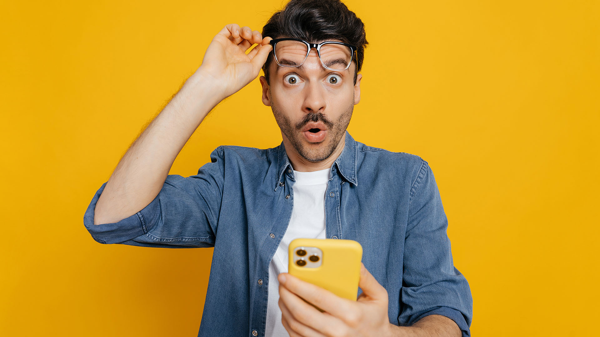 Brunette man in blue jean coat and white shirt looking surprised at his phone. Yellow background.