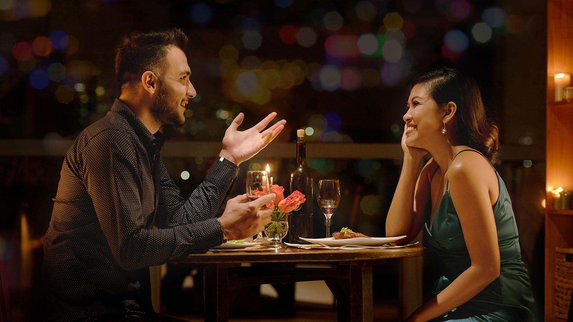 Brunette man and Asian woman at a romantic dinner date. The woman is smiling while looking at the man who is talking.