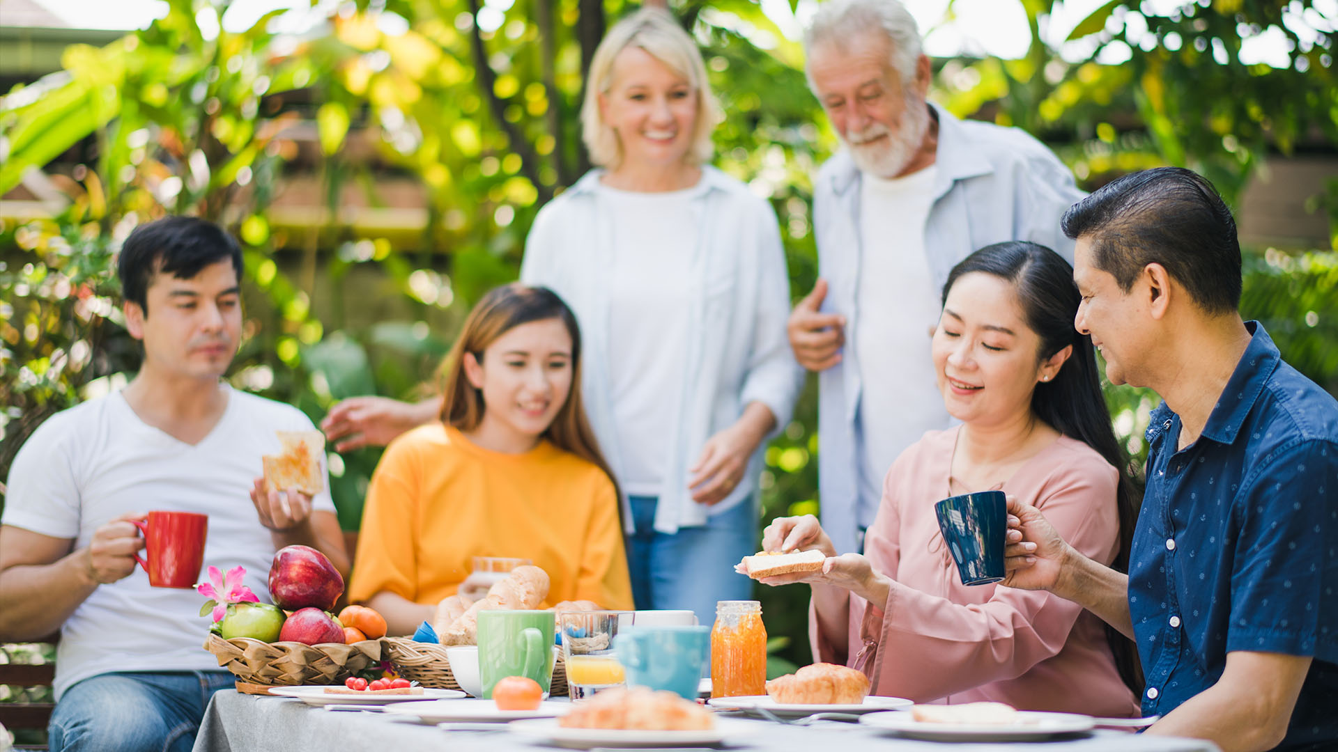 Family of Asian woman and European man sitting down having lunch and smiling while drinking coffee and eating fruit.