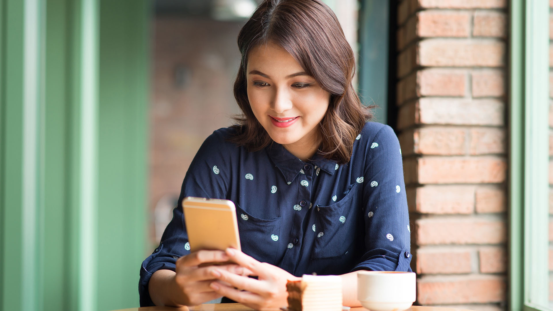 Brunette Asian woman wearing a blue shirt with white dots, sitting down having her coffee while smiling at her phone.