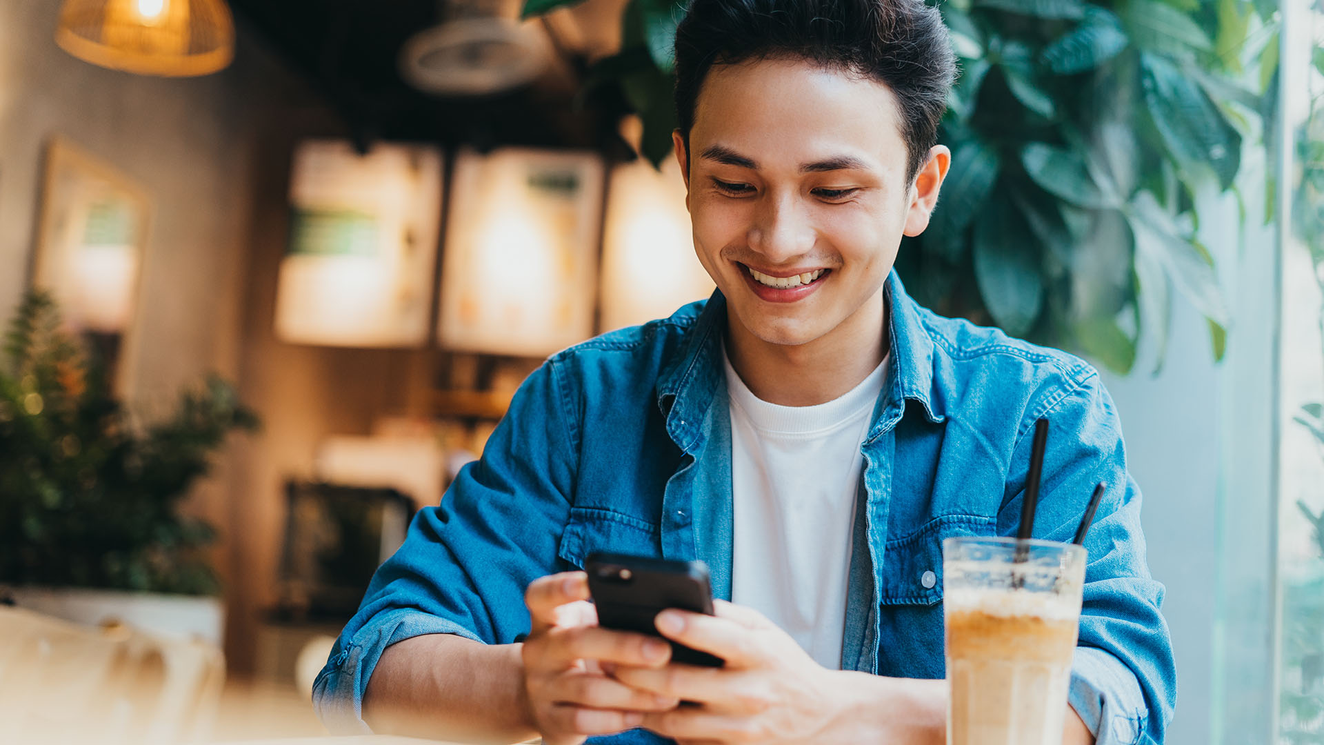 Brunette Asian man wearing a blue jean coat and white shirt, sitting down having his coffee while smiling at his phone.