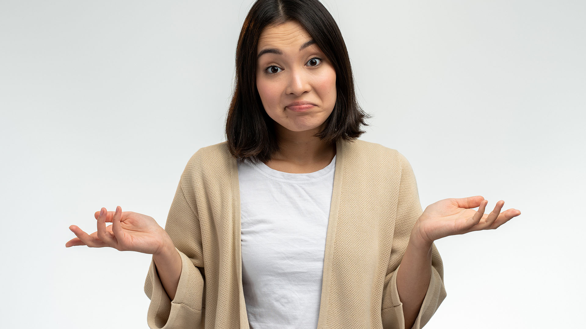 Brunette Asian woman in beige coat and white shirt, confused while shrugging her shoulders with her hands on each side.