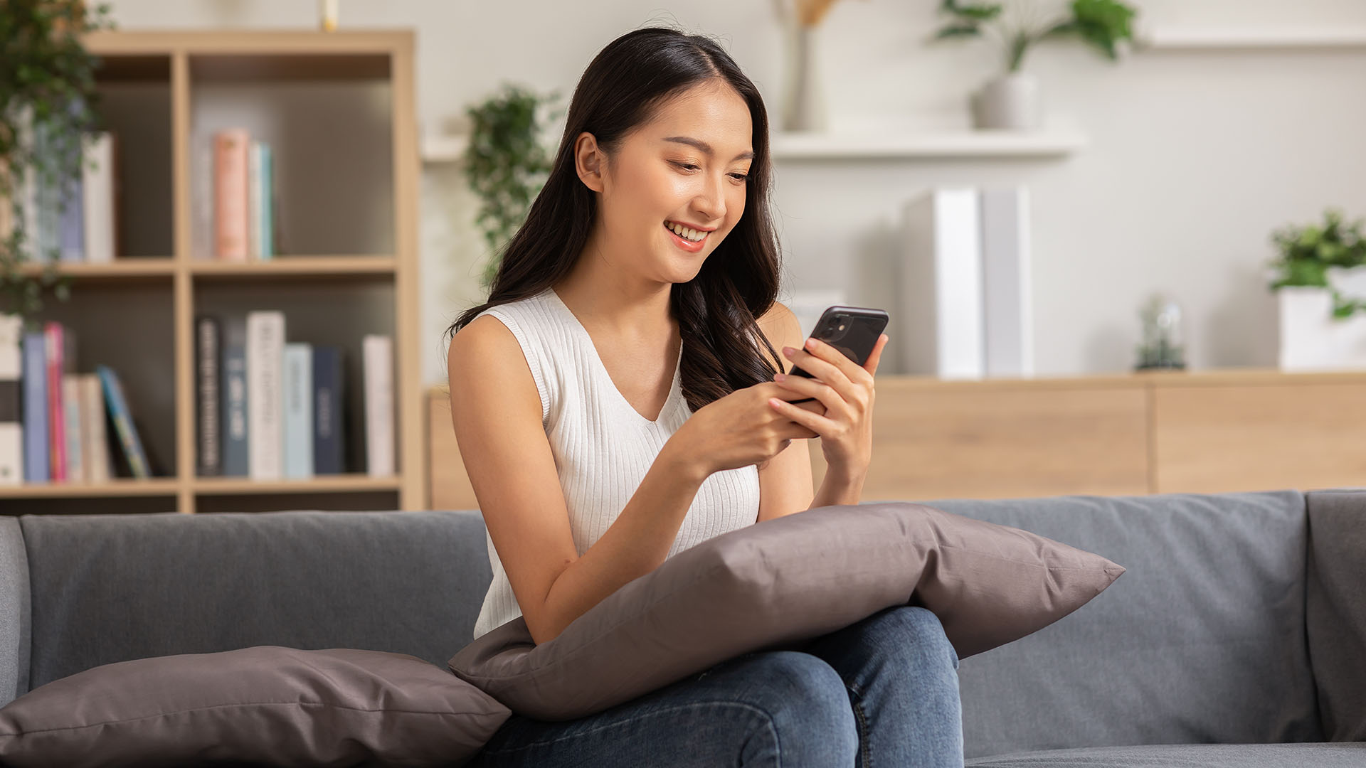 Brunette Asian woman in white shirt and jeans, sitting down at her living room, smiling while looking at her phone.