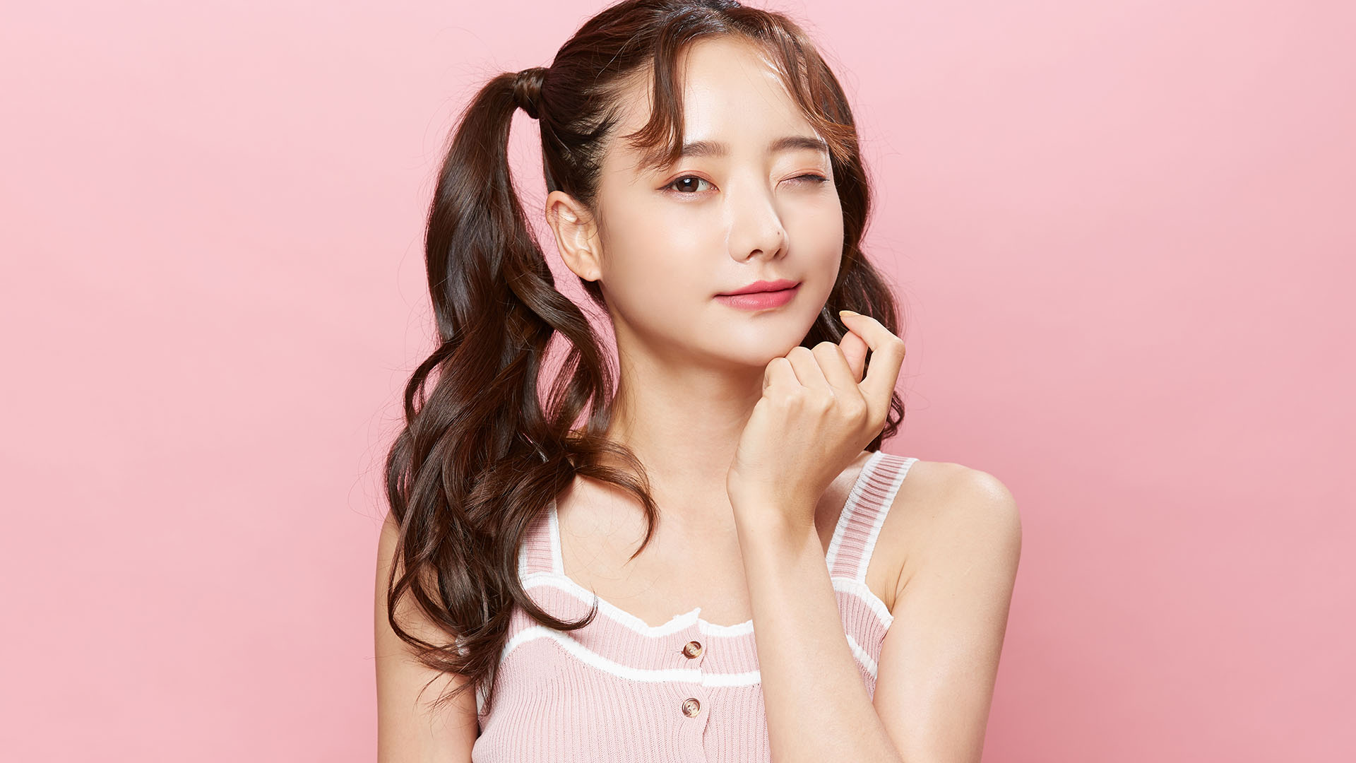 Brunette Asian girl with pigtails, winking at the camera while leaning her chin on her knuckles. Pink background.