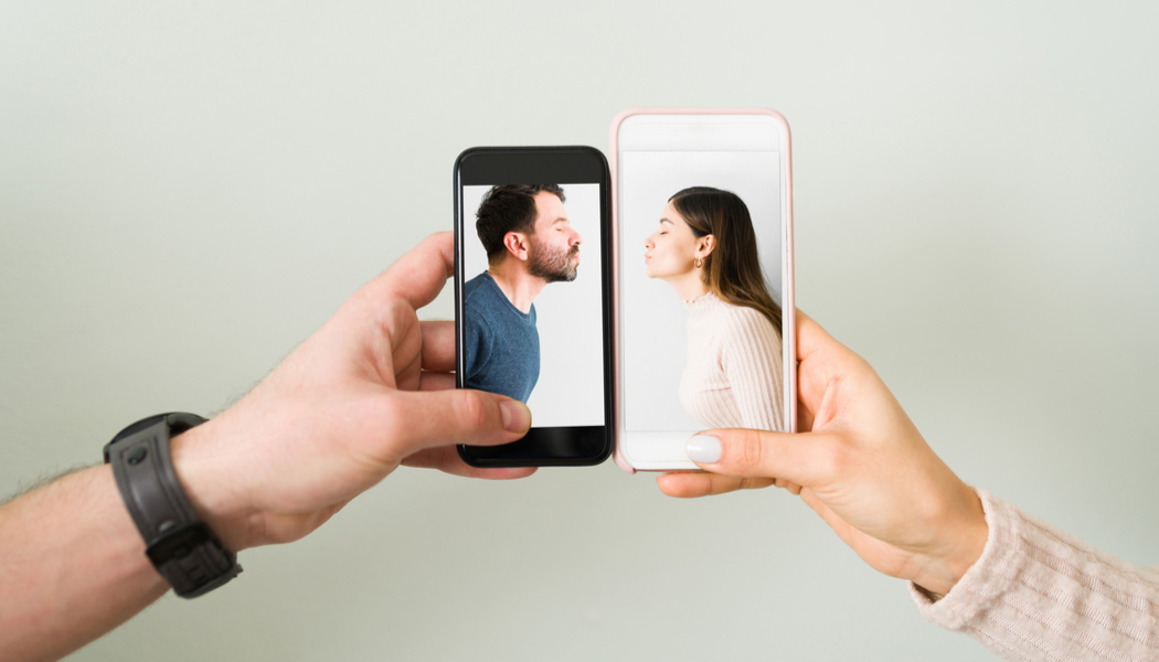 Hands of man and woman holding mobile phones next to each other. The screens show man and woman leaning in for a kiss.