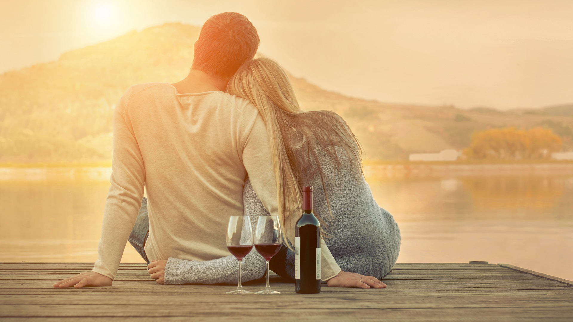 Couple wearing beige and light blue jumpers sitting on the pier with two glasses and a bottle of wine right behind them.