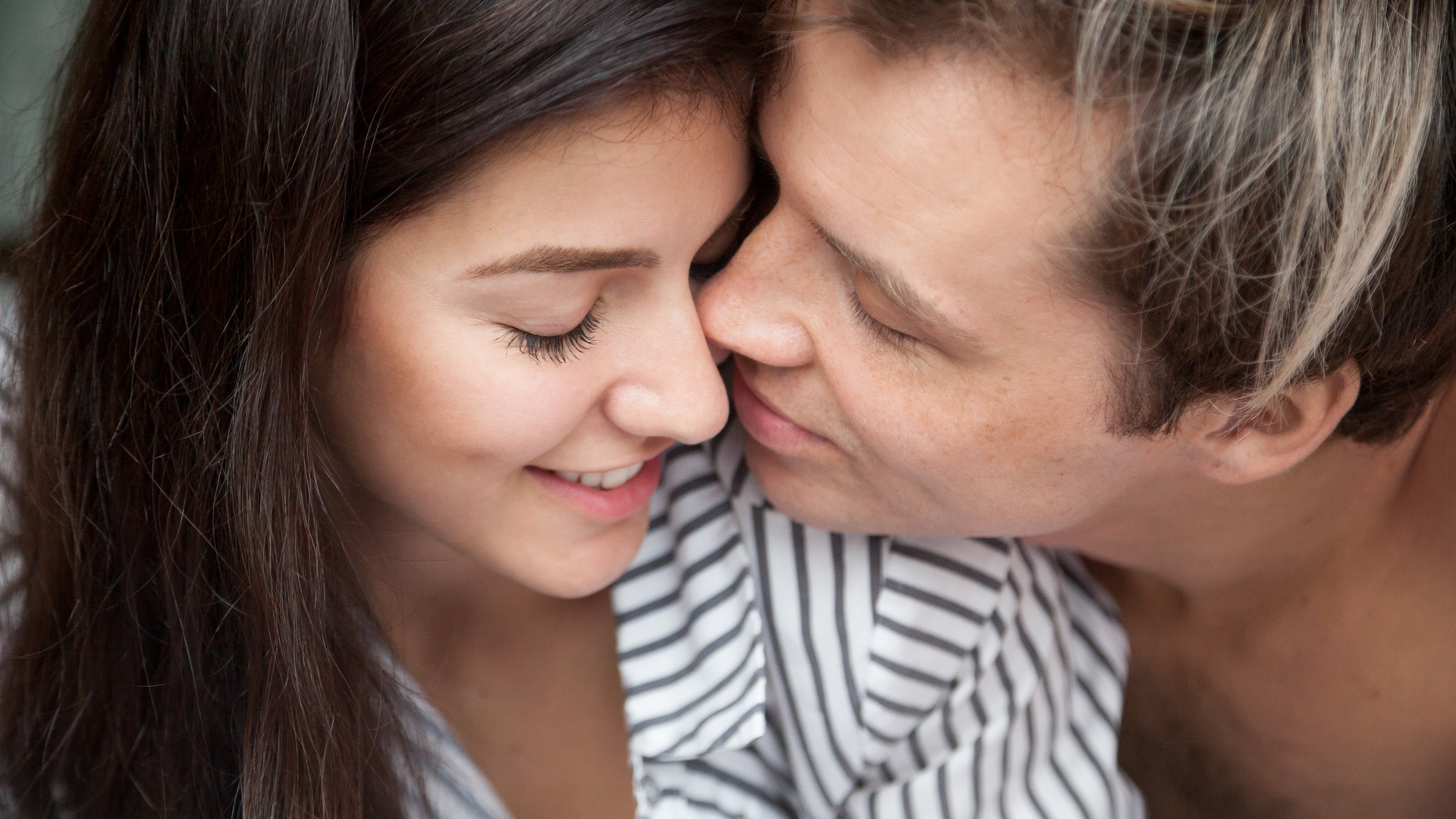 Brunette woman and man with blonde highlights hugging with their eyes closed and their faces close to each other.