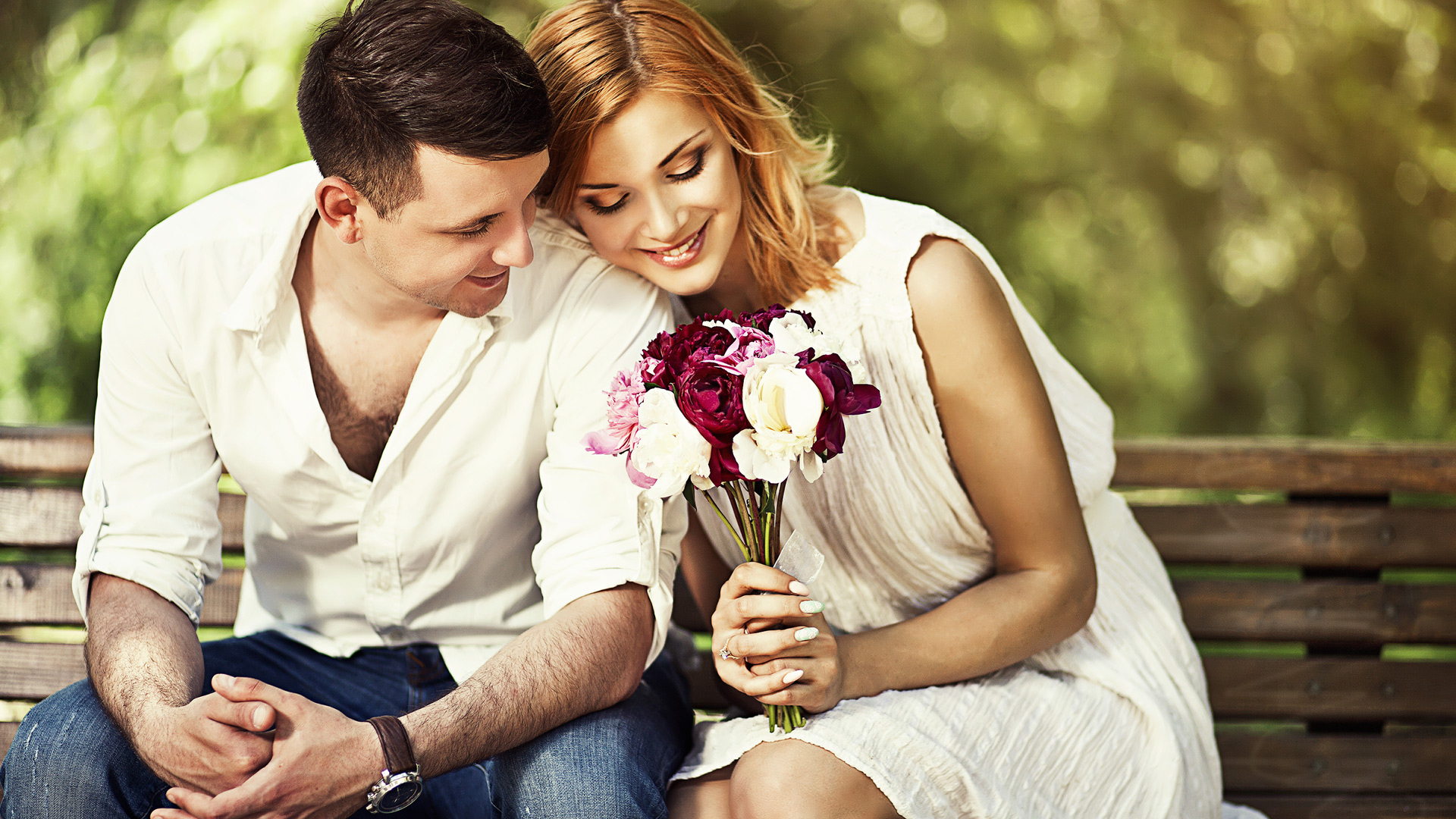 Man in white shirt and woman in white dress sitting next to each other on bench looking at the flowers the woman is holding.
